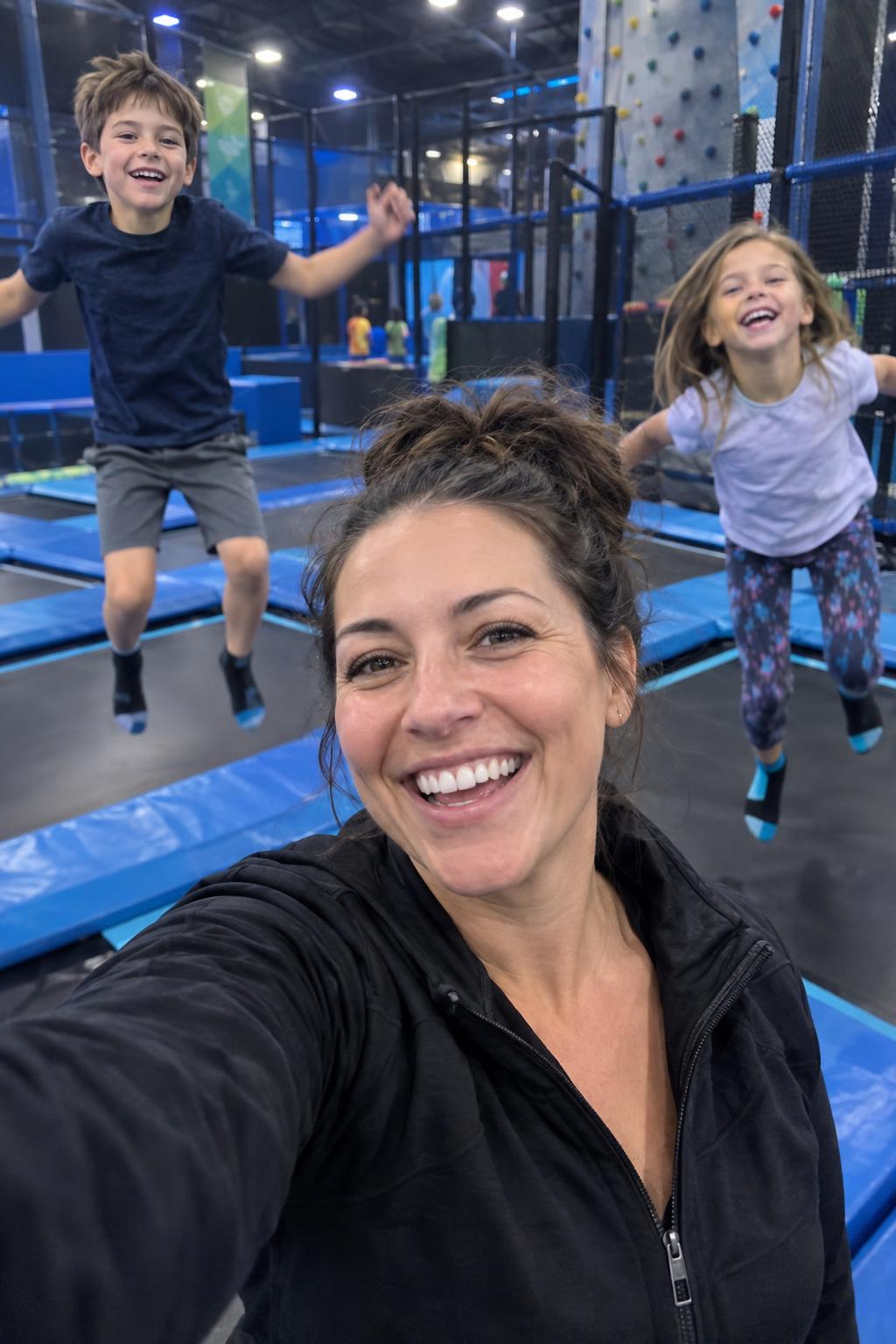 Mom filming a selfie video at an indoor family fun center while her kids run and jump behind her